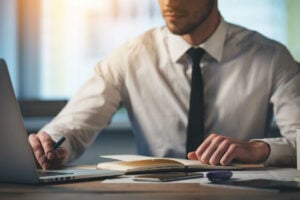 Midsection of man sitting at computer, professionally dressed in a professional setting, the frame cuts off his face from the nose up
