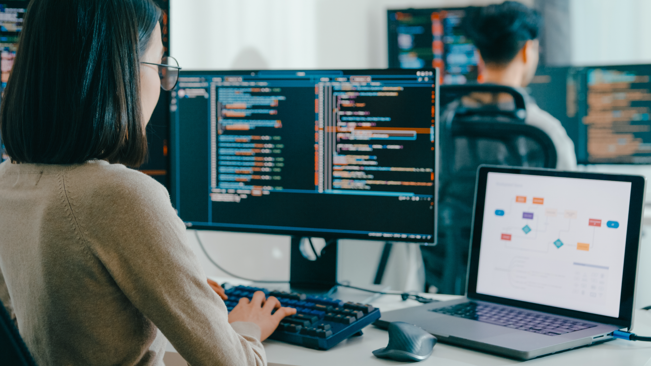  Female developer with glasses typing code on dual monitors at office workstation with a colleague working in the background