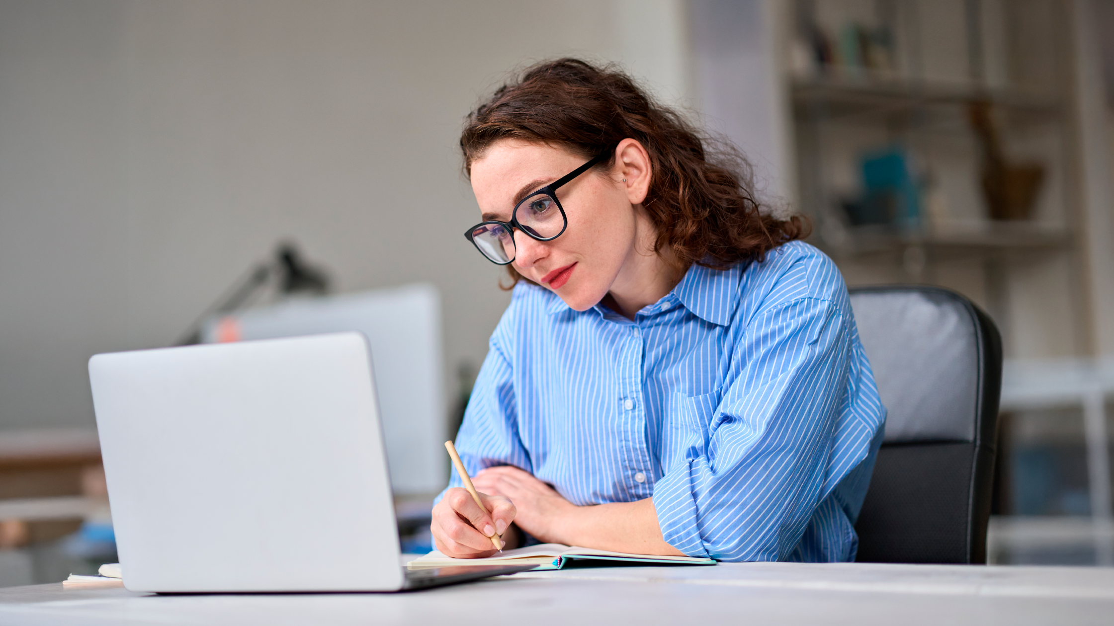 Young professional woman with glasses writing notes beside laptop in modern office