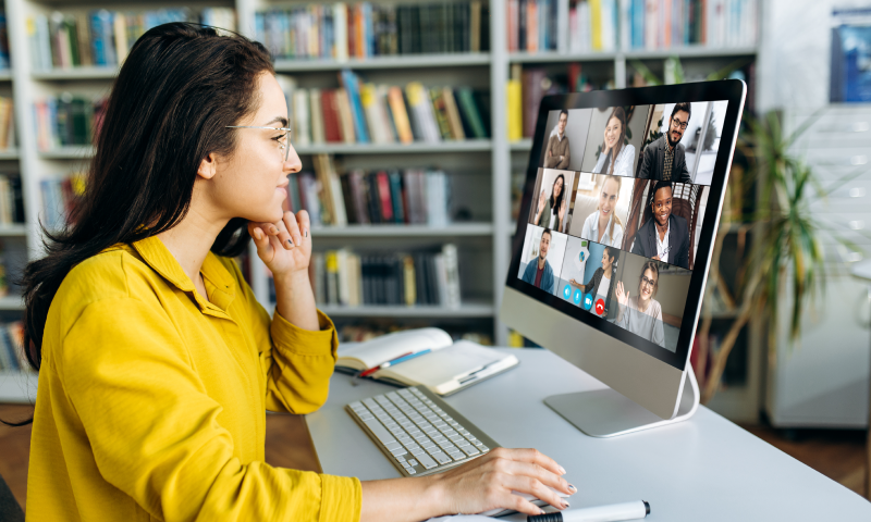 Woman in yellow shirt attending video conference with diverse group on desktop computer in library