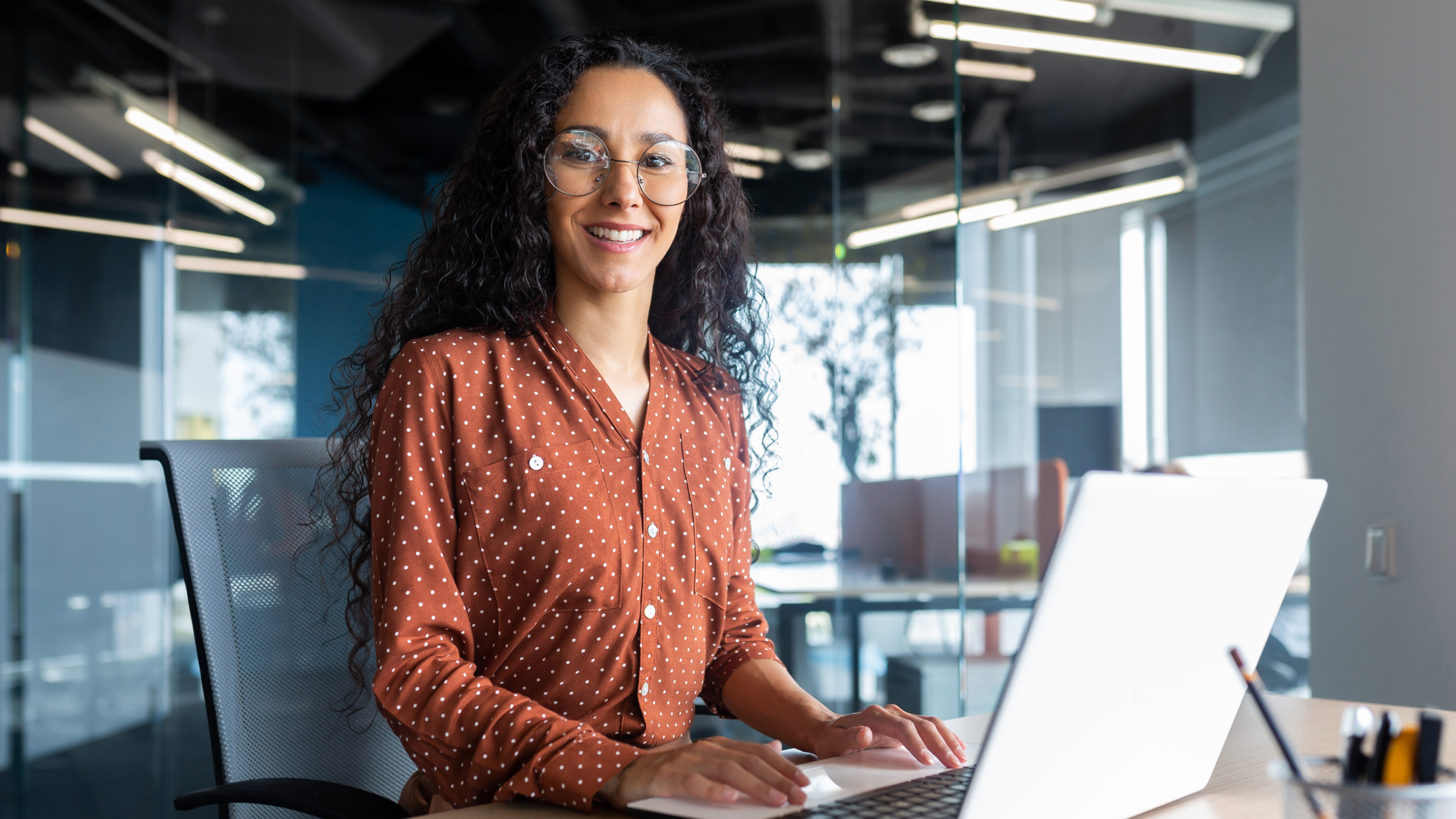 Smiling professional woman with curly hair and glasses working on laptop in modern glass office