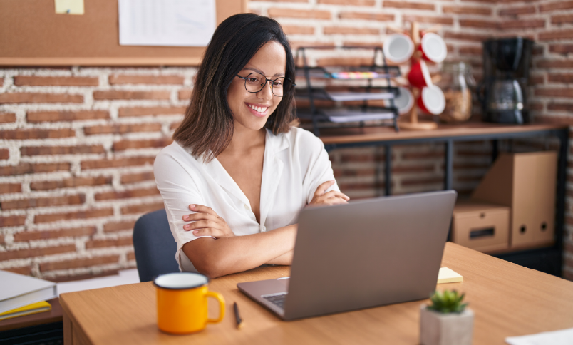 Smiling woman with glasses working on laptop at office desk with coffee mug and small plant