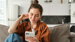 Young Gen Z woman smiling while scrolling smartphone on couch at home