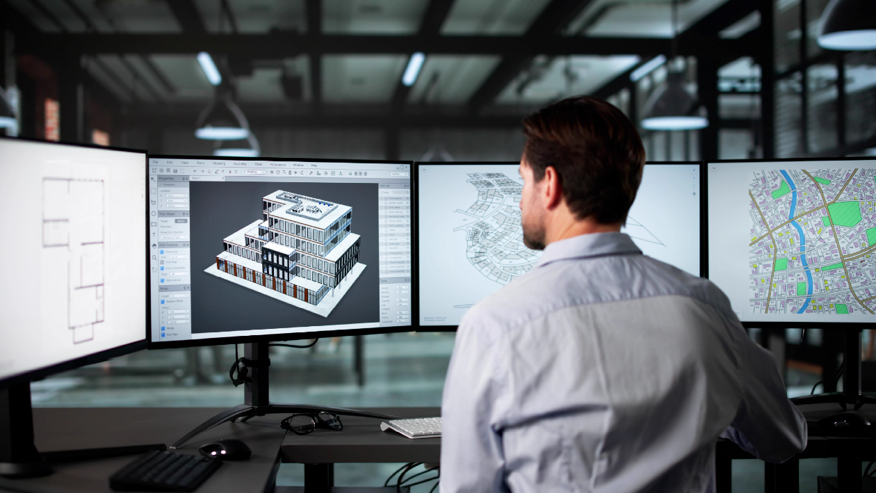 Male engineer reviewing 3D building model on multi-monitor workstation with CAD software and city map