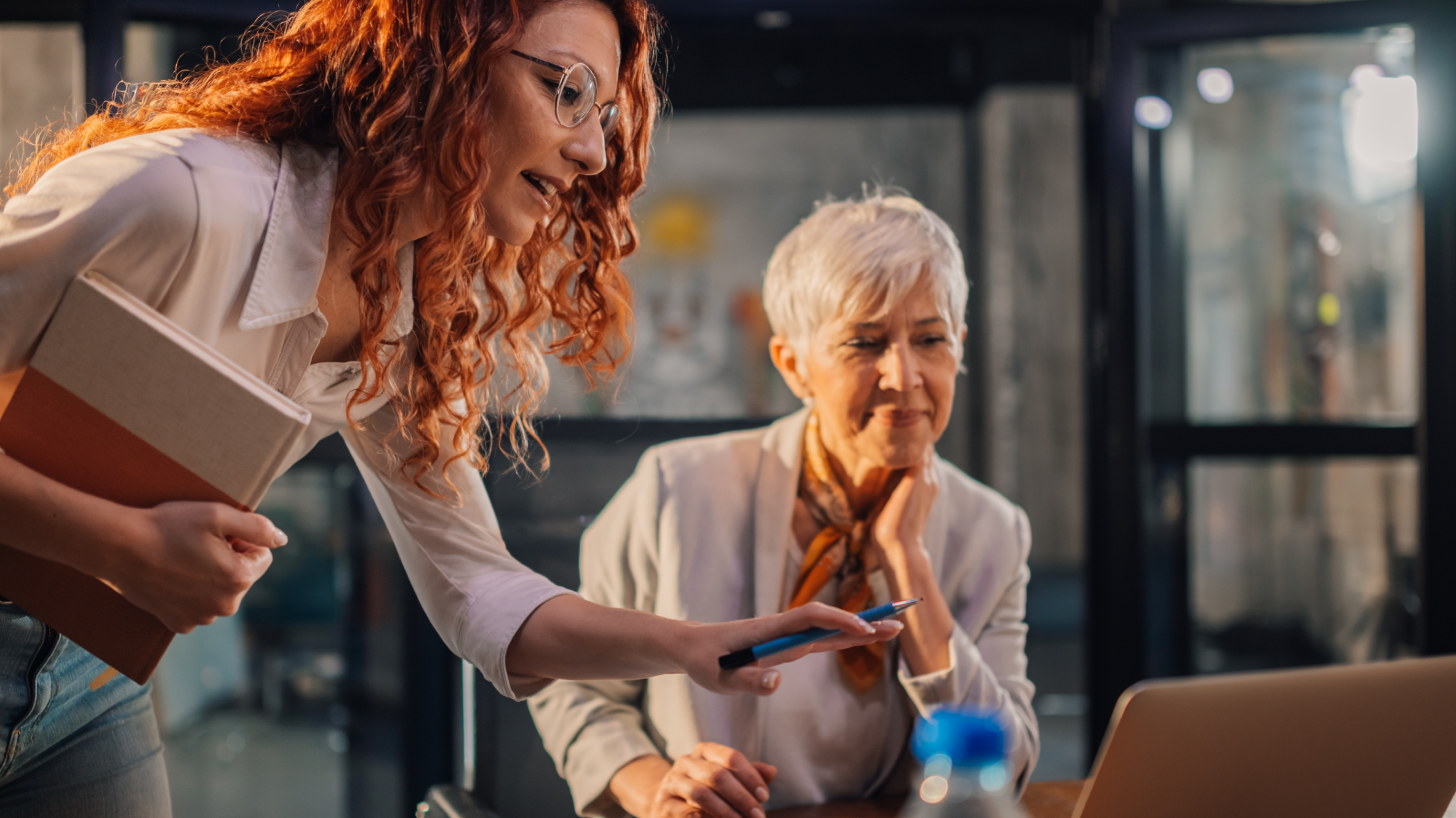 Young professional woman leaning over desk pointing at laptop screen with senior female colleague in office