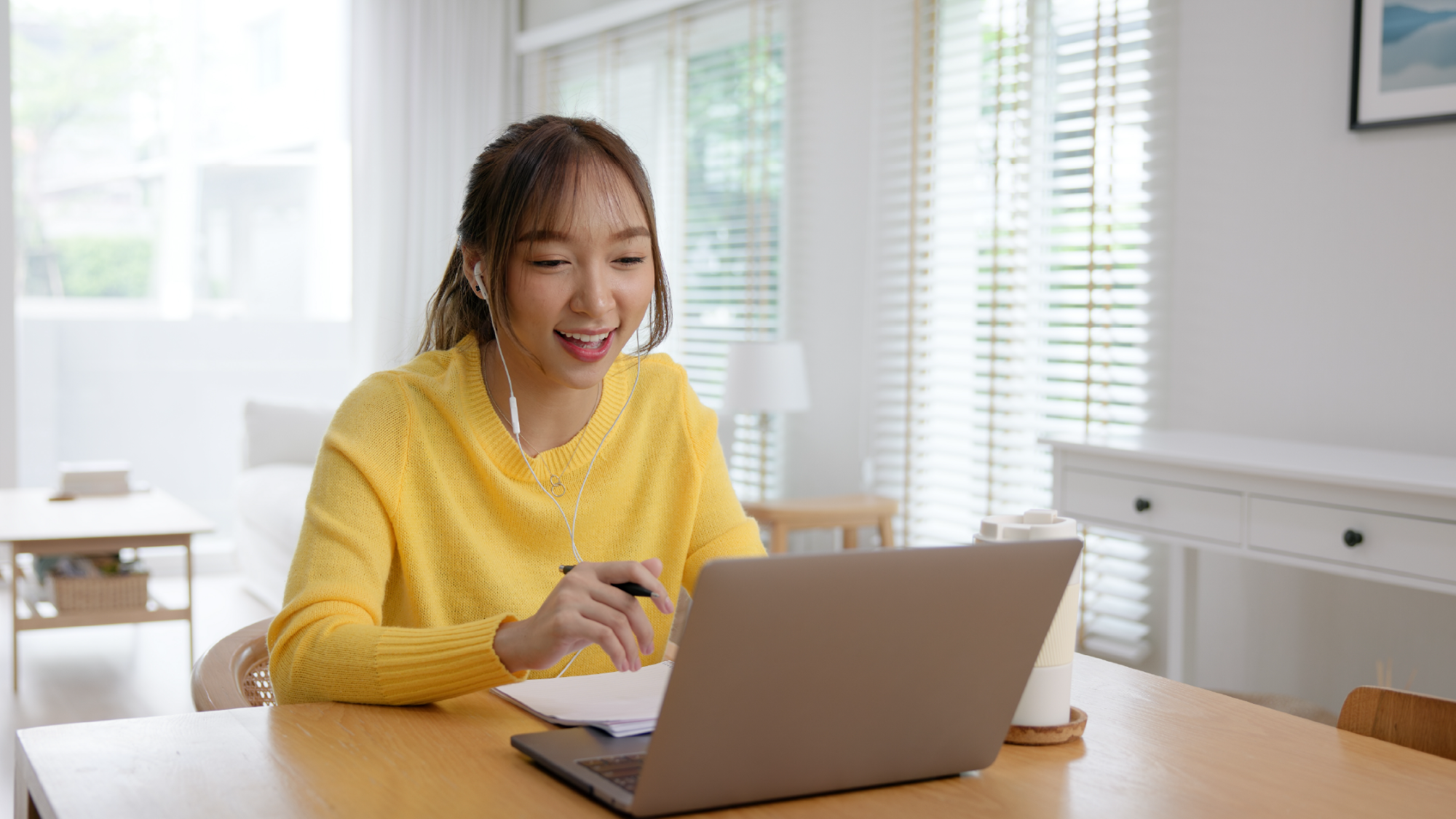 Young woman in yellow sweater smiling at laptop with earbuds during online class at home